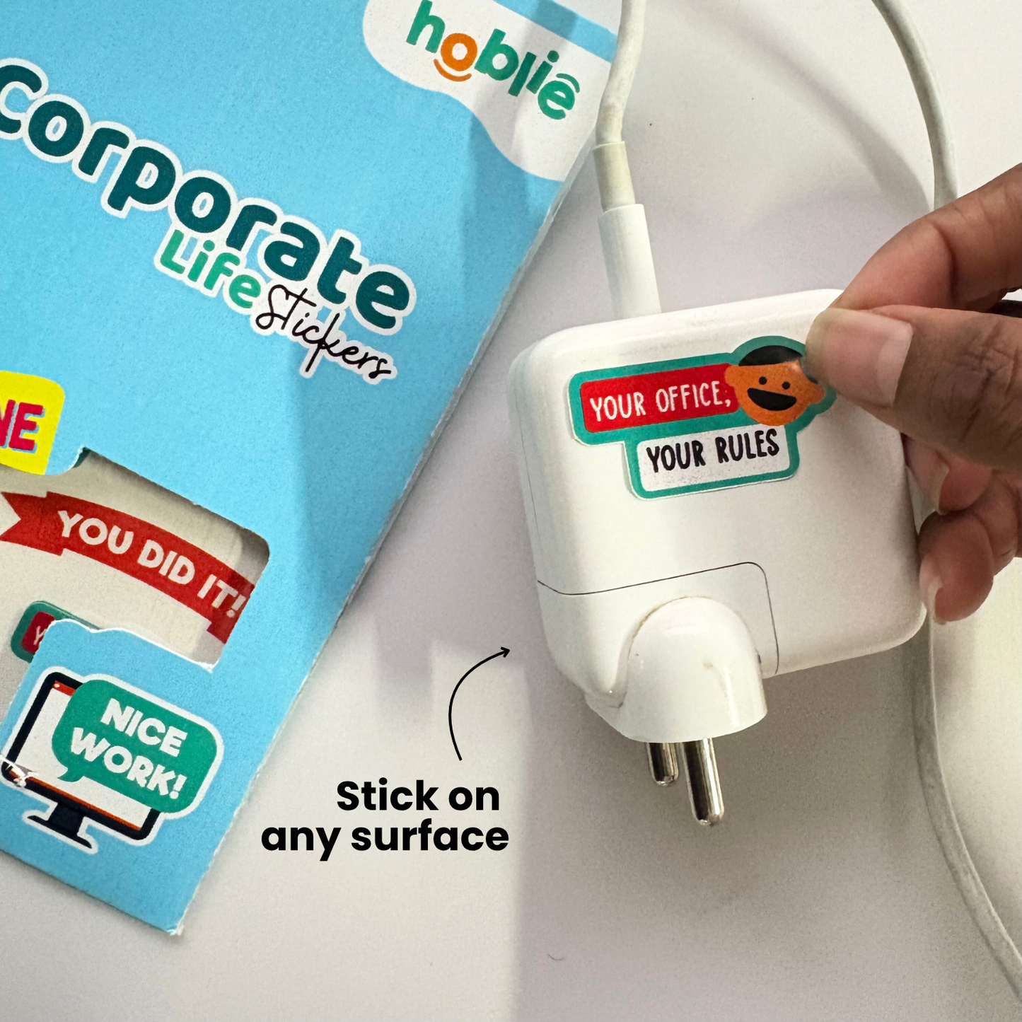 Hand holding a white plug with a sticker on it, next to a box of stickers with text and graphics.